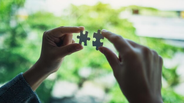 Closeup image of a woman holding and putting a piece of white jigsaw puzzle together with green nature background