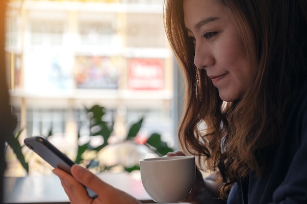 Closeup image of a woman holding , using and looking at smart phone while drinking coffee in cafe