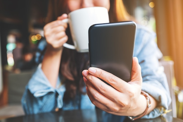 Closeup image of a woman holding  and using mobile phone while drinking coffee