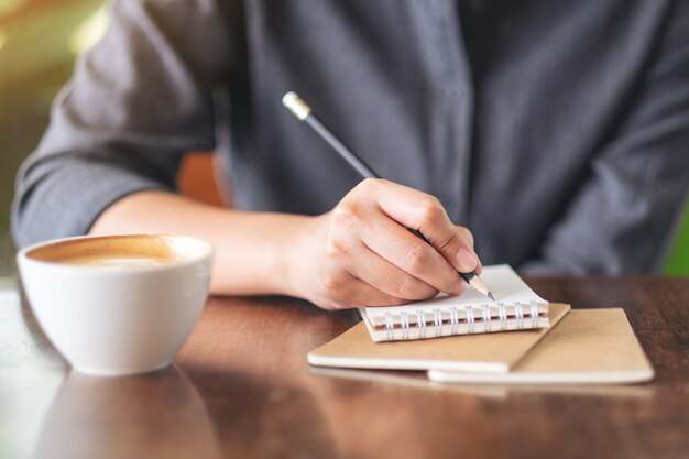 Closeup image of a woman writing on blank notebook with coffee cup on table in cafe