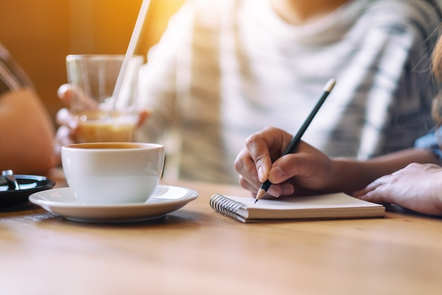 Closeup image of a woman writing on a notebook while drinking coffee with friends