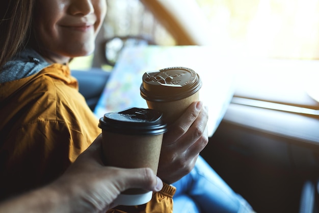 Closeup image of a young woman holding a map and clinking coffee cup with friend while riding in the car