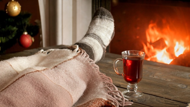 Closeup image of young woman in woolen socks lying under blanket next to burning fireplace with glass of mulled wine