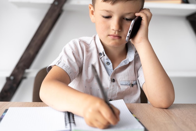 Closeup of a little student talking on a cell phone A boy sits at a desk talks on a smartphone and makes notes in a notebook Technology and education concept