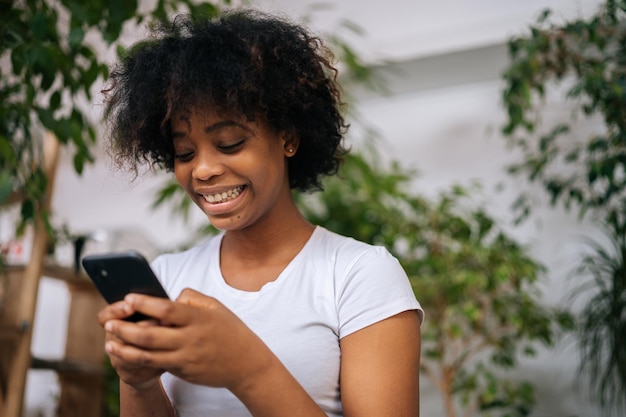 Closeup lowangle view face of cheerful curly AfricanAmerican young woman holding mobile phone in hands enjoying web surfing communicating in social network