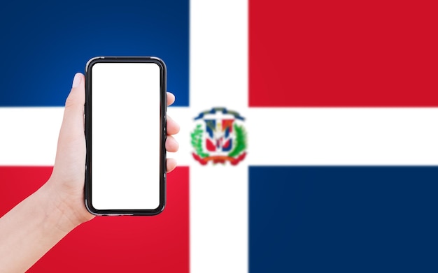 Closeup of male hand holding smartphone with blank on screen on background of blurred flag of Dominican Republic