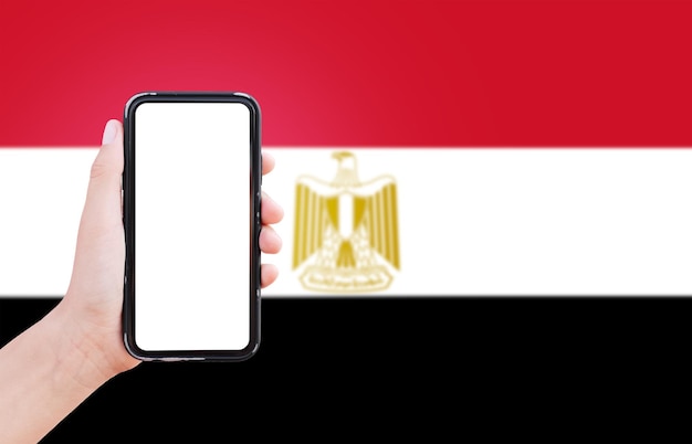 Closeup of male hand holding smartphone with blank on screen on background of blurred flag of Egypt