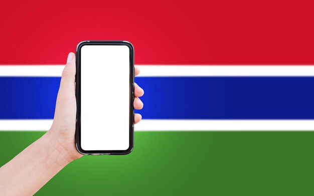 Closeup of male hand holding smartphone with blank on screen on background of blurred flag of The Gambia