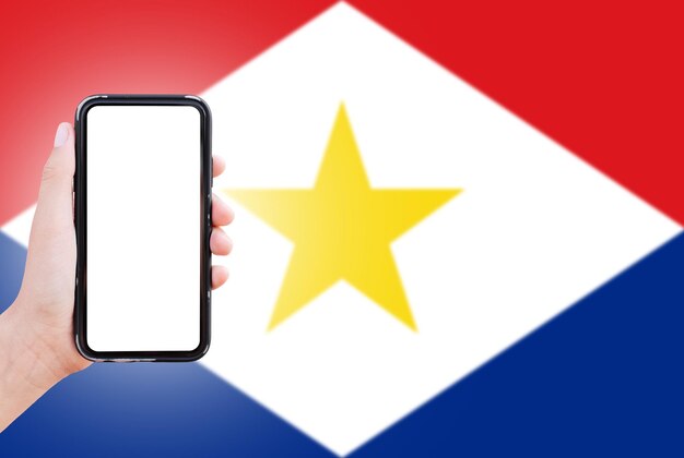 Closeup of male hand holding smartphone with blank on screen on background of blurred flag of Saba