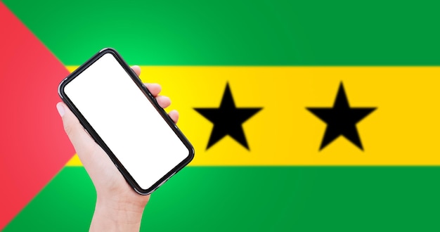 Closeup of male hand holding smartphone with blank on screen on background of blurred flag of Sao Tome and Principe