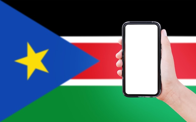 Closeup of male hand holding smartphone with blank on screen on background of blurred flag of South Sudan