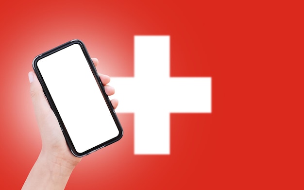 Closeup of male hand holding smartphone with blank on screen on background of blurred flag of Switzerland