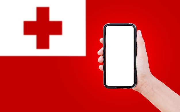 Closeup of male hand holding smartphone with blank on screen on background of blurred flag of Tonga