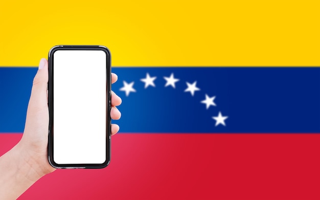 Closeup of male hand holding smartphone with blank on screen on background of blurred flag of Venezuela