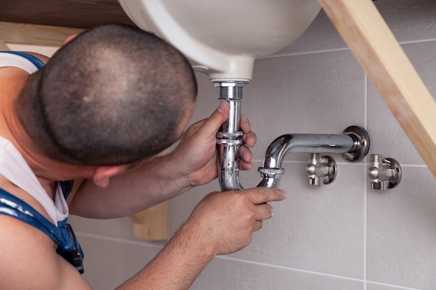 Closeup male plumber in blue denim uniform fixing sink in bathroom with tile wall