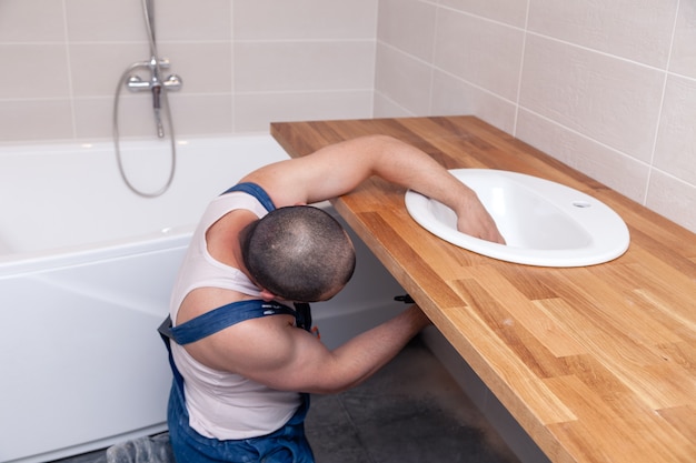 Closeup male plumber worker in blue denim uniform, overalls, fixing sink in bathroom with tile wall. Professional plumbing repair service, installation water pipes, man mounted sewer drain