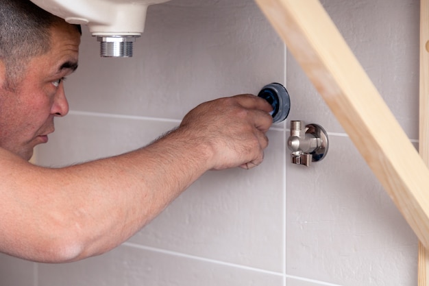 Closeup male plumber worker in blue denim uniform, overalls, fixing sink in bathroom with tile wall. Professional plumbing repair service, installation water pipes, man mounted sewer drain