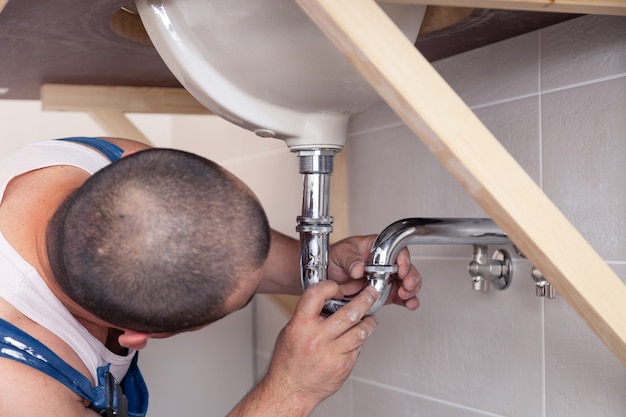 Closeup male plumber worker in blue denim uniform, overalls, fixing sink in bathroom with tile wall. Professional plumbing repair service, installation water pipes, man mounted sewer drain