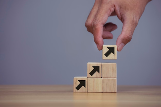 Closeup man hand arranging wood cube stacking as step stair with arrow up symbol