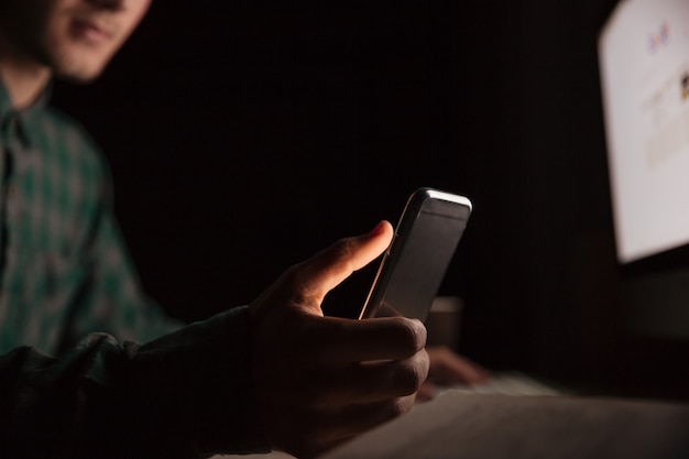 Closeup of man sitting at the table and using cell phone in the dark room