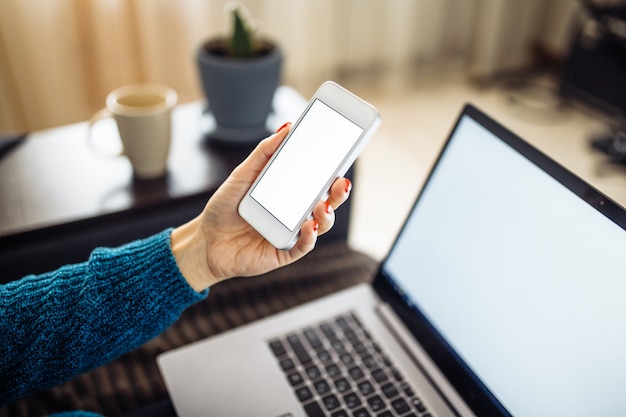 Closeup of a mobile cell phone of a young woman staying home and leadling business from distance on a freelance basis. Laptop, cup and flower in the room on the background.