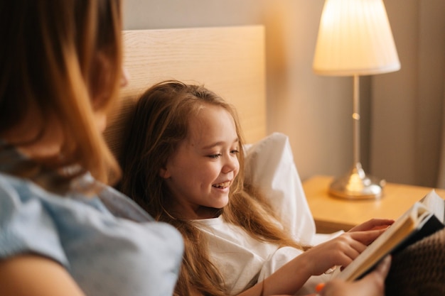 Closeup of mother and cheerful adorable daughter reading together children book before going to sleep while lying in bed