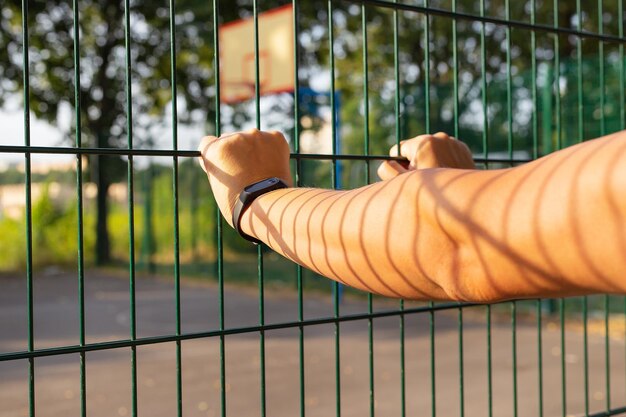 Photo closeup of a person s hand gripping a green metal fence with a blurred background of trees and a basketball court represents strength security and boundaries