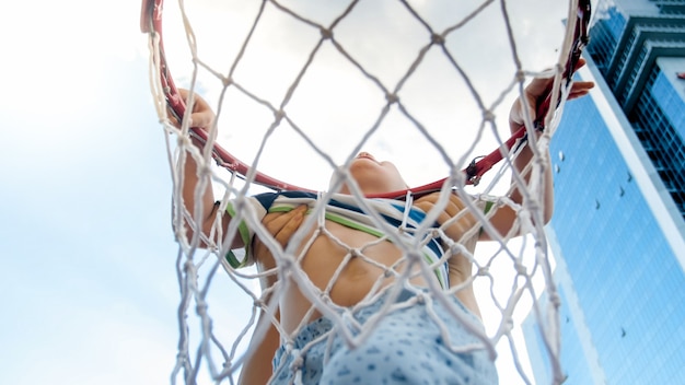 Closeup photo of active 3 years old toddler boy holding on basketball net ring. Concept of active and sporty children. Health of new generation kids