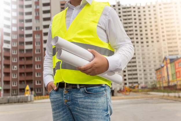 Closeup photo of architect in safety vest posing with blueprints at new buildings