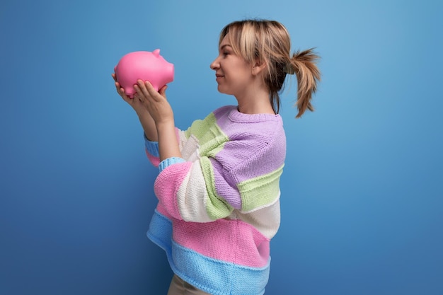 Closeup photo of an attractive blond woman in a striped sweater with a piggy bank of banknotes on a