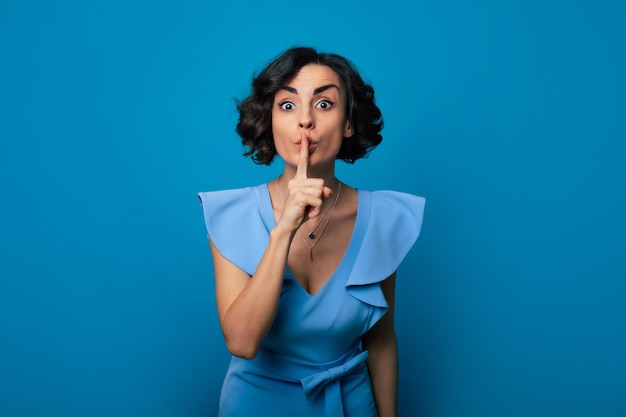 Closeup photo of a beautiful girl in a blue dress who is looking in the camera while pressing her index finger to her lips and isolated on blue background