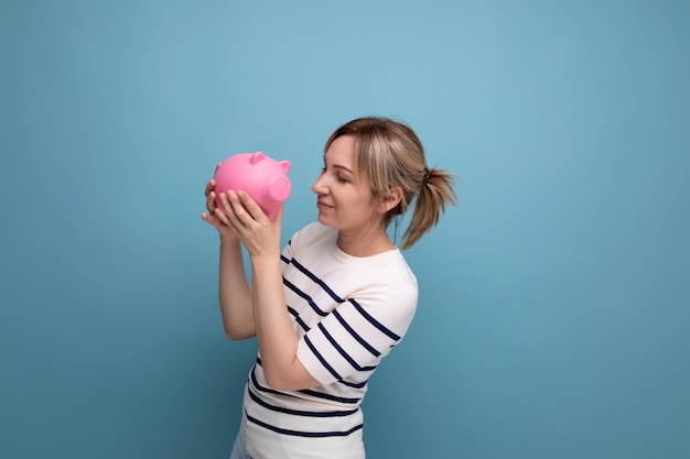 Closeup photo of blond charming young woman in casual outfit thoughtfully holding a piggy bank with