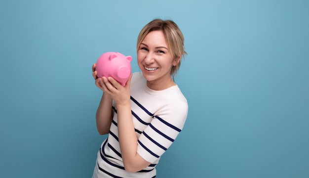 Closeup photo of blond charming young woman in casual outfit thoughtfully holding a piggy bank with