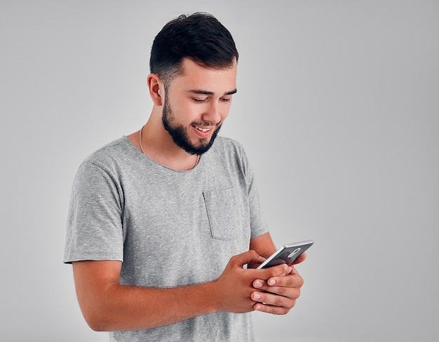 Closeup photo of casually dressed student standing isolated on gray background looking at screen of smart phone, browsing web pages and smiling while chatting with friends