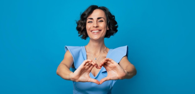 Closeup photo of a cheerful cute woman in a long blue dress who is looking in the camera and smiling while showing a heart with her hands isolated on blue background