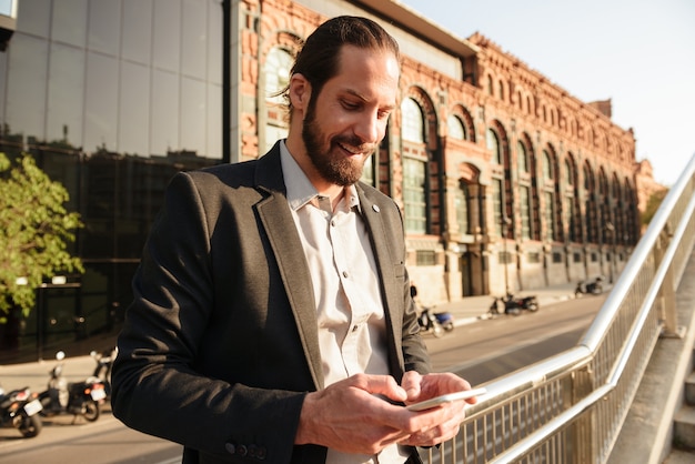 Closeup photo of european handsome man 30s in formal suit using smartphone, while standing in front of office building or business centre