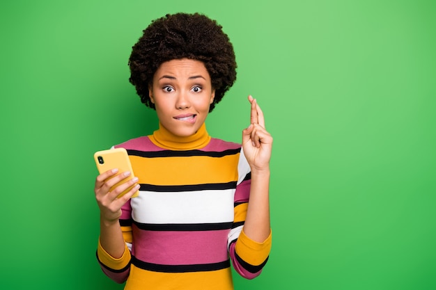 Closeup photo of funny dark skin wavy lady hold telephone fingers crossed wait startup investment results bite lips worried wear casual striped jumper 