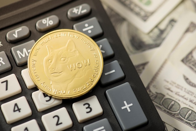 Closeup photo of gold coin with dogecoin symbol on calculator and hundred dollar banknotes