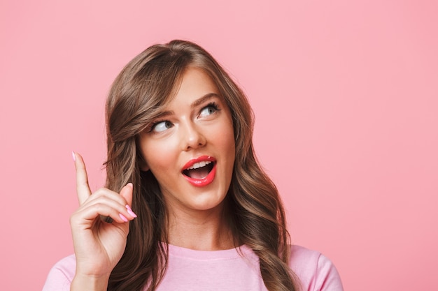 Closeup photo of happy cute woman with long curly brown hair looking upward and pointing finger at copyspace, isolated over pink background