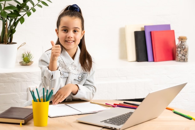 Closeup photo of little pretty pupil school lady talk skype notebook online lesson video call sit desk use earphones distance greet classmates wave arm quarantine study living room indoors