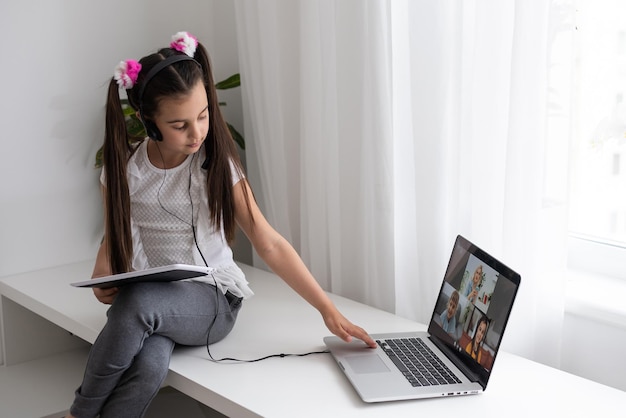 Closeup photo of little pretty pupil school lady talk skype notebook online lesson video call sit desk use earphones distance greet classmates wave arm quarantine study living room indoors.