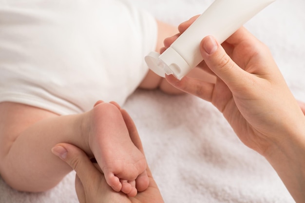 Closeup photo of newborn laying on stomach and mother's hands holding baby's foot and white cream tube on isolated white textile background