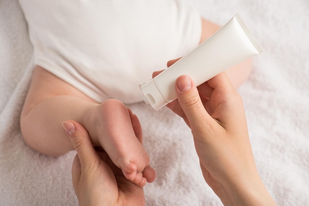 Closeup photo of newborn laying on stomach and mother's hands holding baby's leg and white lotion tube on isolated white blanket background