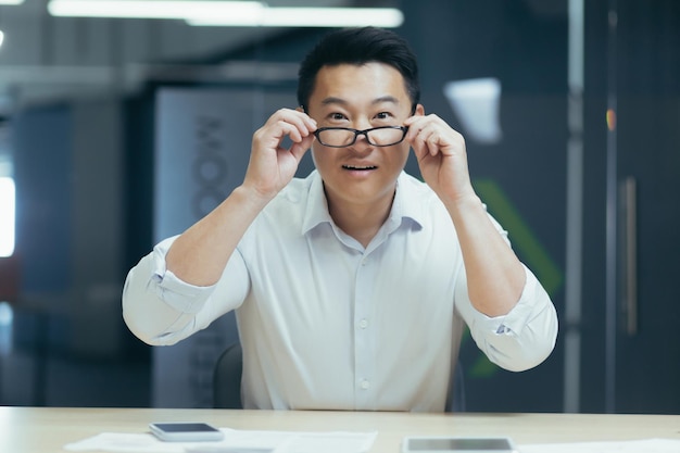 Closeup photo portrait of a young smiling asian man sitting in the office at the desk and holding