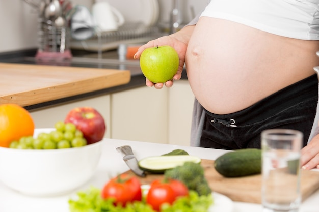 Closeup photo of pregnant woman posing with green apple on kitchen