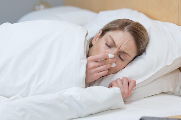 Closeup photo of a sick woman lying under a blanket in bed at home has a runny nose and cold