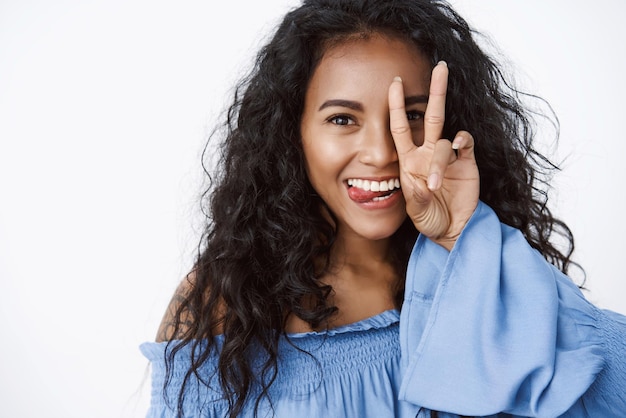 Closeup playful and feminine coquettish africanamerican curlyhaired female in blue blouse silly showing tongue make peace goodwill sign over face having fun and joking with positive emotions