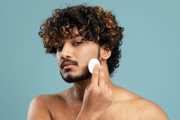 Closeup portrait of attractive Indian man removing makeup using cotton disk, cleaning face.Skin care