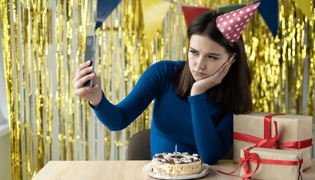 Closeup portrait of a bored birthday girl sitting with a phone in her hands waiting for a call or video online Distance party at home One for a holiday Cake and gifts on the woman's table