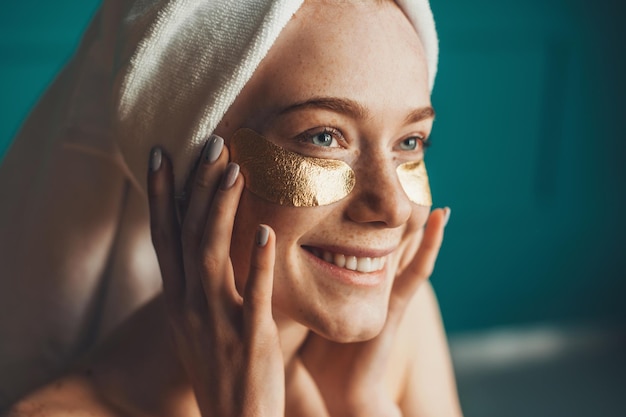 Closeup portrait of a caucasian freckled woman wearing white towel on head with collagen golden patc